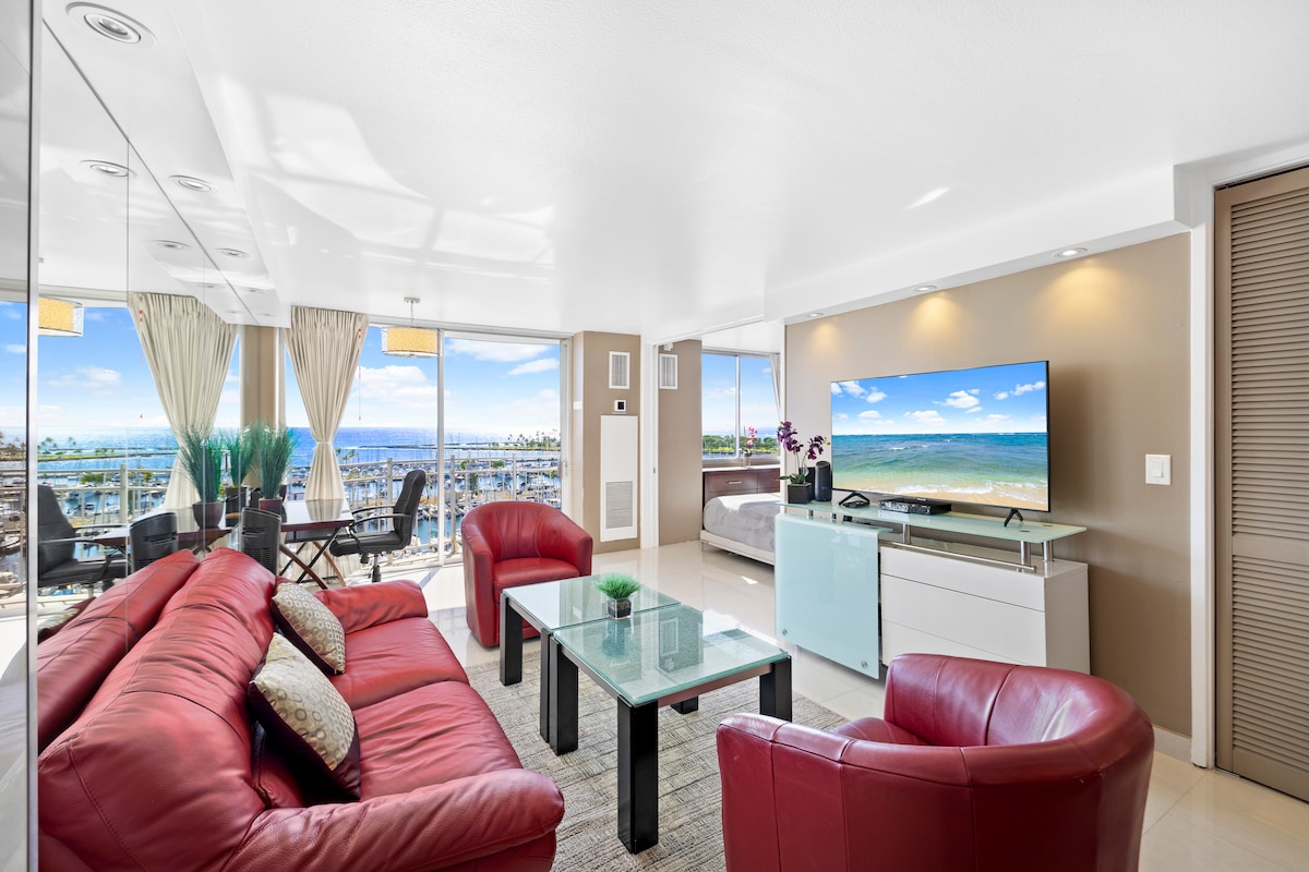 A modern living area features a large television mounted on the wall above a sleek white console. Comfortable red leather sofas surround a glass coffee table. Floor-to-ceiling windows provide a view of the ocean, while light floods the space through sheer curtains.