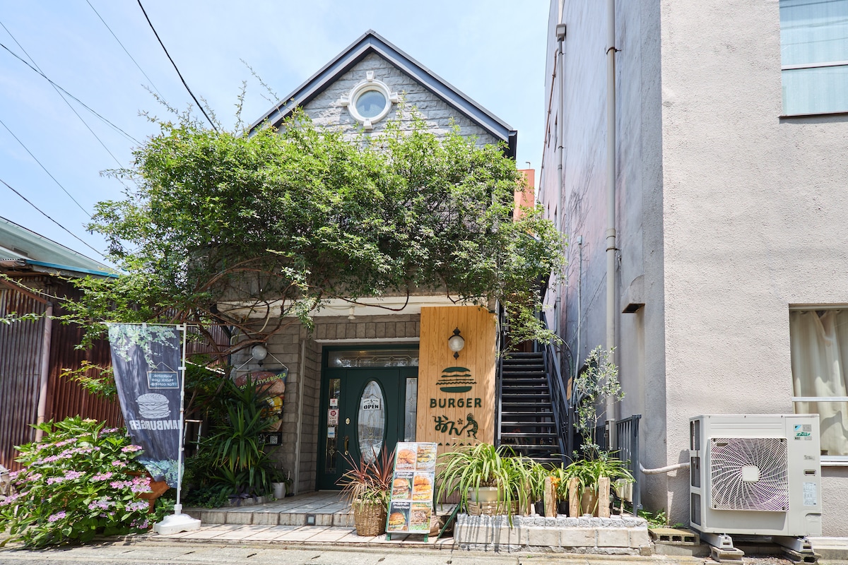 The entrance to the restaurant features a rustic wooden sign displaying the name and offers a glimpse of lush greenery above. Steps lead to a welcoming doorway, with various menus displayed nearby. Surrounding plants contribute to a tranquil setting, complementing the building's charm.
