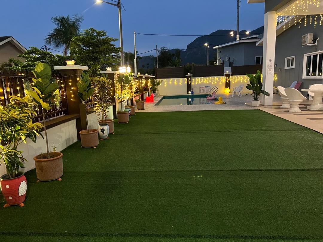 A well-maintained outdoor area features a lush green lawn surrounded by potted plants. Soft lighting creates a gentle ambiance, illuminating the private swimming pool in the background. The space is designed for gatherings, offering ample room for relaxation and leisure activities.