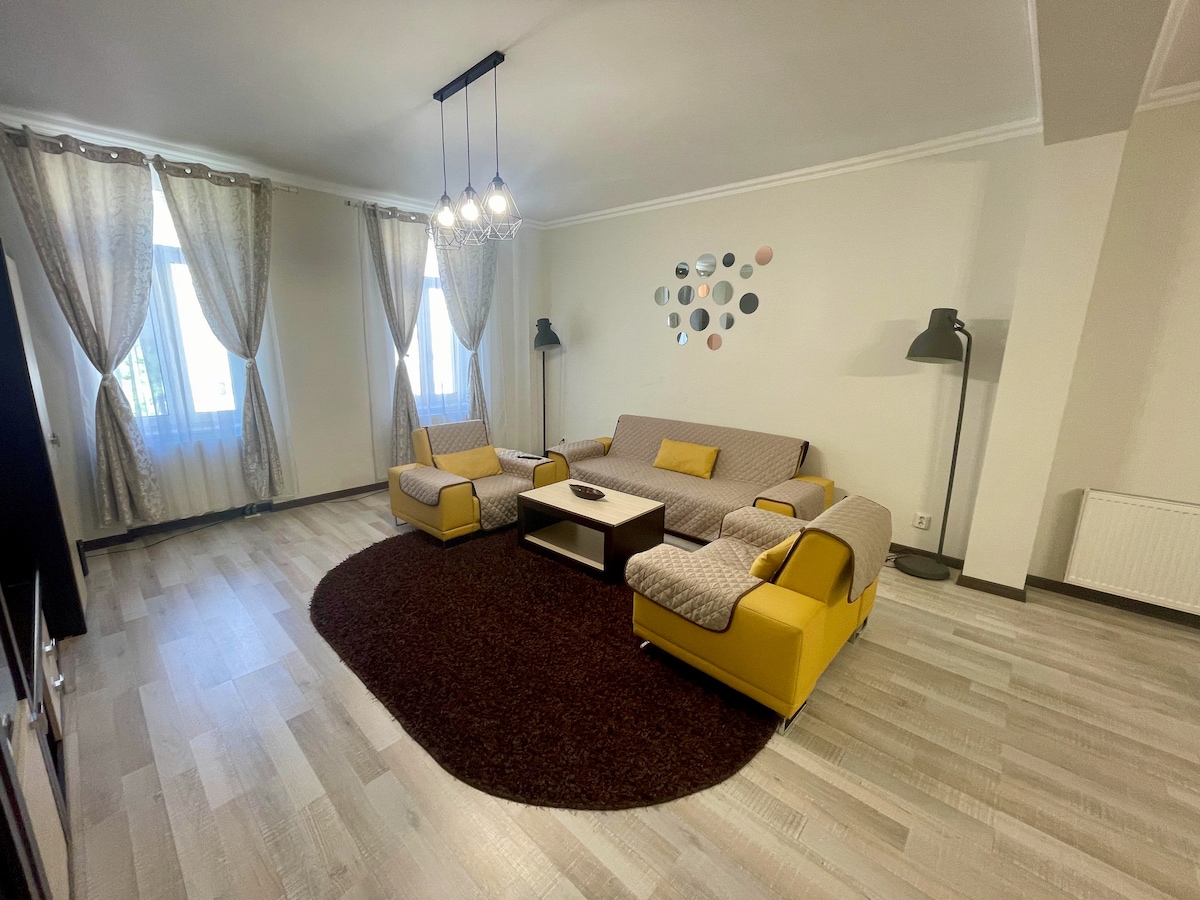 A spacious living room features comfortable yellow and grey sofas arranged around a coffee table on a large brown area rug. Natural light filters through sheer curtains at the large windows, complemented by a modern floor lamp and decorative wall art.