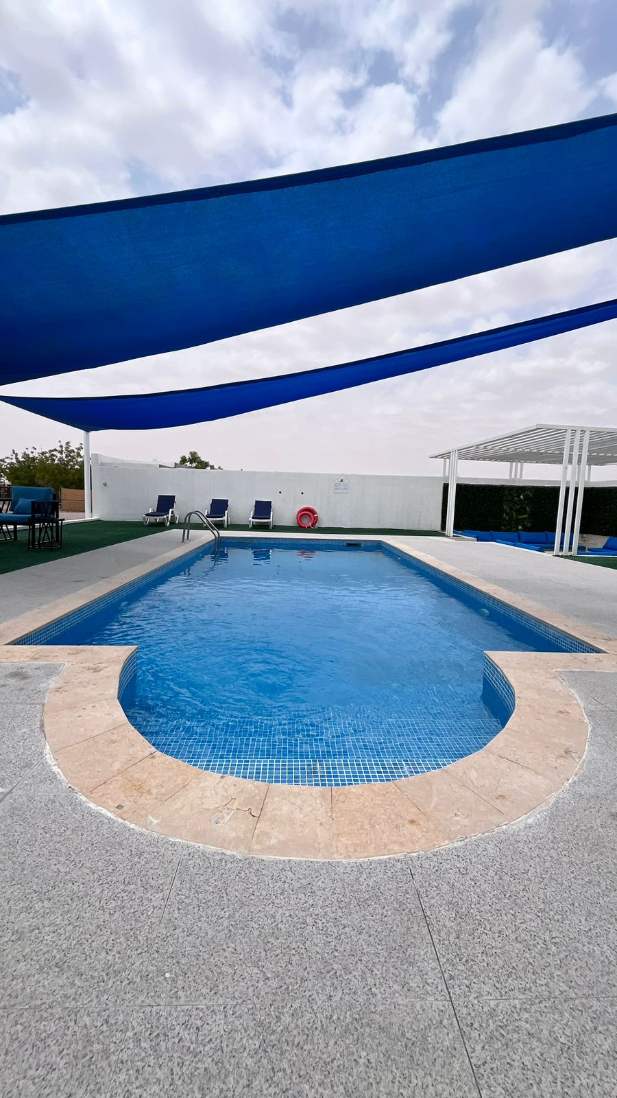 A swimming pool is surrounded by a stone border, with blue sunshade sails overhead. Lounge chairs are positioned nearby, offering a place to relax. Green landscaping and a white wall create a serene backdrop, enhancing the inviting atmosphere of the outdoor space.