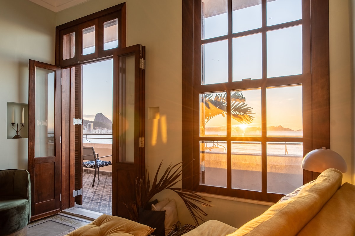 Large windows fill the room with natural light, showcasing a view of the sunrise. The double doors open wide, revealing a balcony that overlooks the beach. A cozy seating area with a yellow sofa is positioned nearby, complemented by indoor plants.