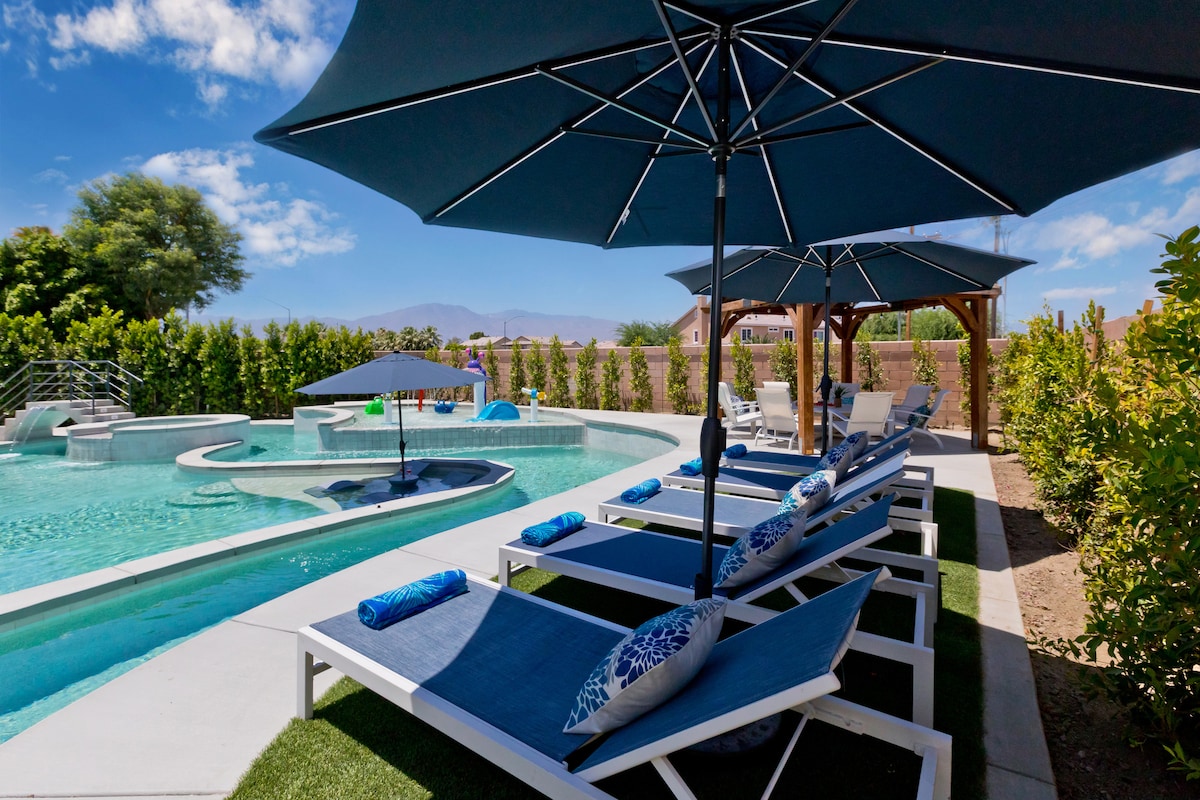Sun loungers with blue cushions are arranged along the edge of a large lazy river pool, featuring clear water and shaded by large umbrellas. Lush greenery surrounds the pool, enhancing the serene environment.