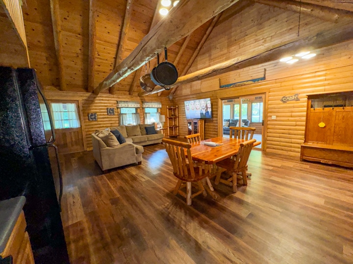 A spacious living area features vaulted wooden ceilings with exposed beams. A large dining table, crafted from logs, is placed in the center, surrounded by matching chairs. A comfortable couch sits nearby, with large windows allowing natural light to illuminate the room.