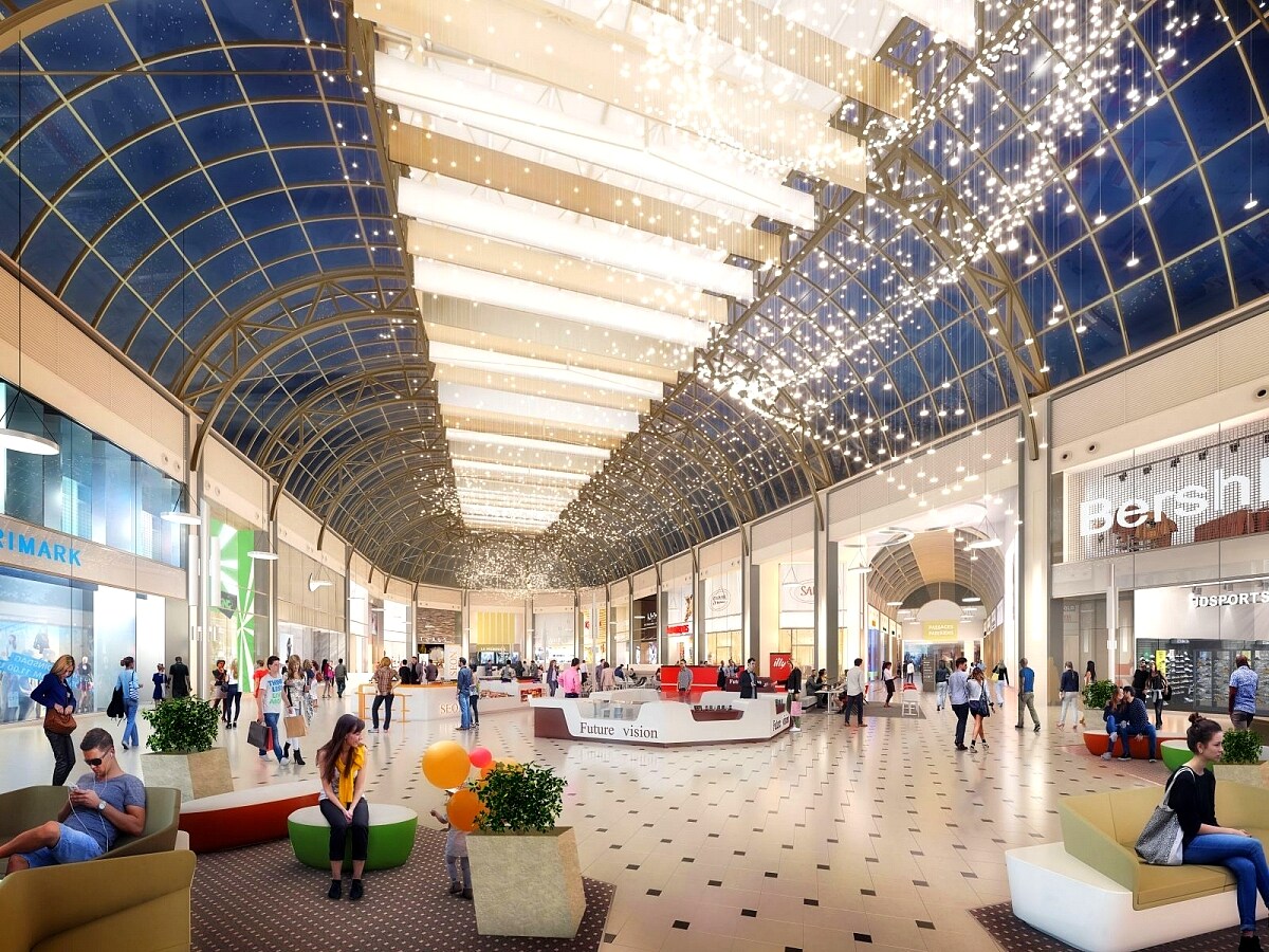 An indoor shopping area features a spacious, modern design with a high glass roof. String lights twinkle overhead while visitors sit and engage in conversation. Storefronts line the sides, providing a variety of shopping options. Potted plants add greenery to the environment.