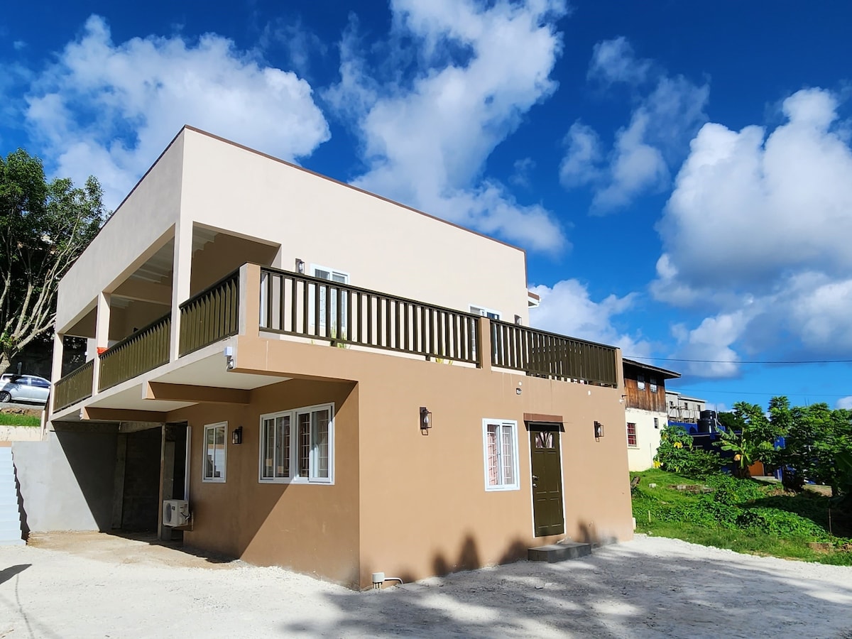 A modern two-story building is set against a clear blue sky filled with scattered clouds. The structure features a wide balcony with railing, and large windows providing ample natural light. The lower level showcases an entrance and a dusted gravel driveway.