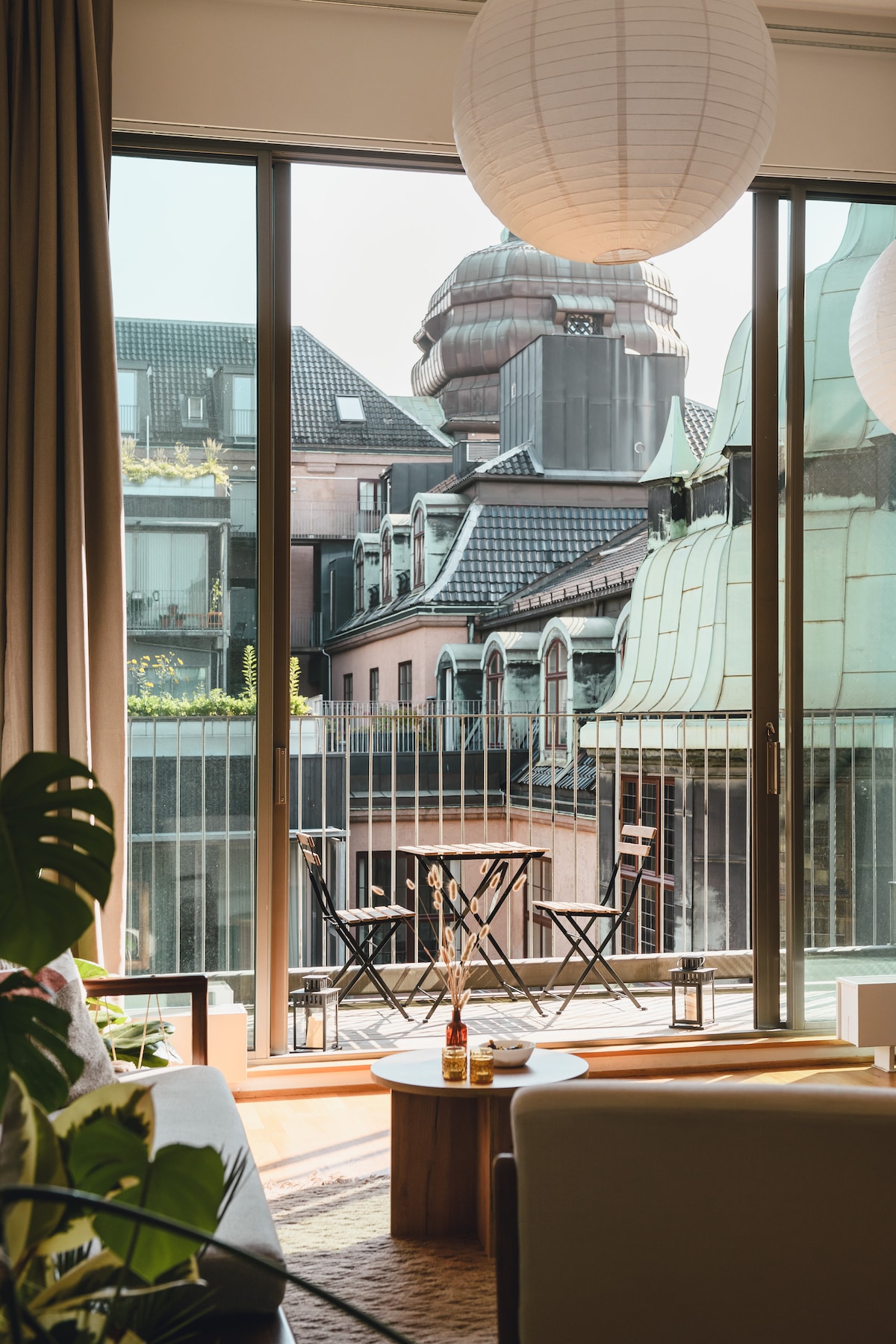 Large glass doors open to a private balcony, revealing views of the tranquil courtyard and surrounding rooftops. A small table with two chairs is positioned on the balcony, while sunlight creates a bright atmosphere in the living area, complemented by decorative pendant lights.
