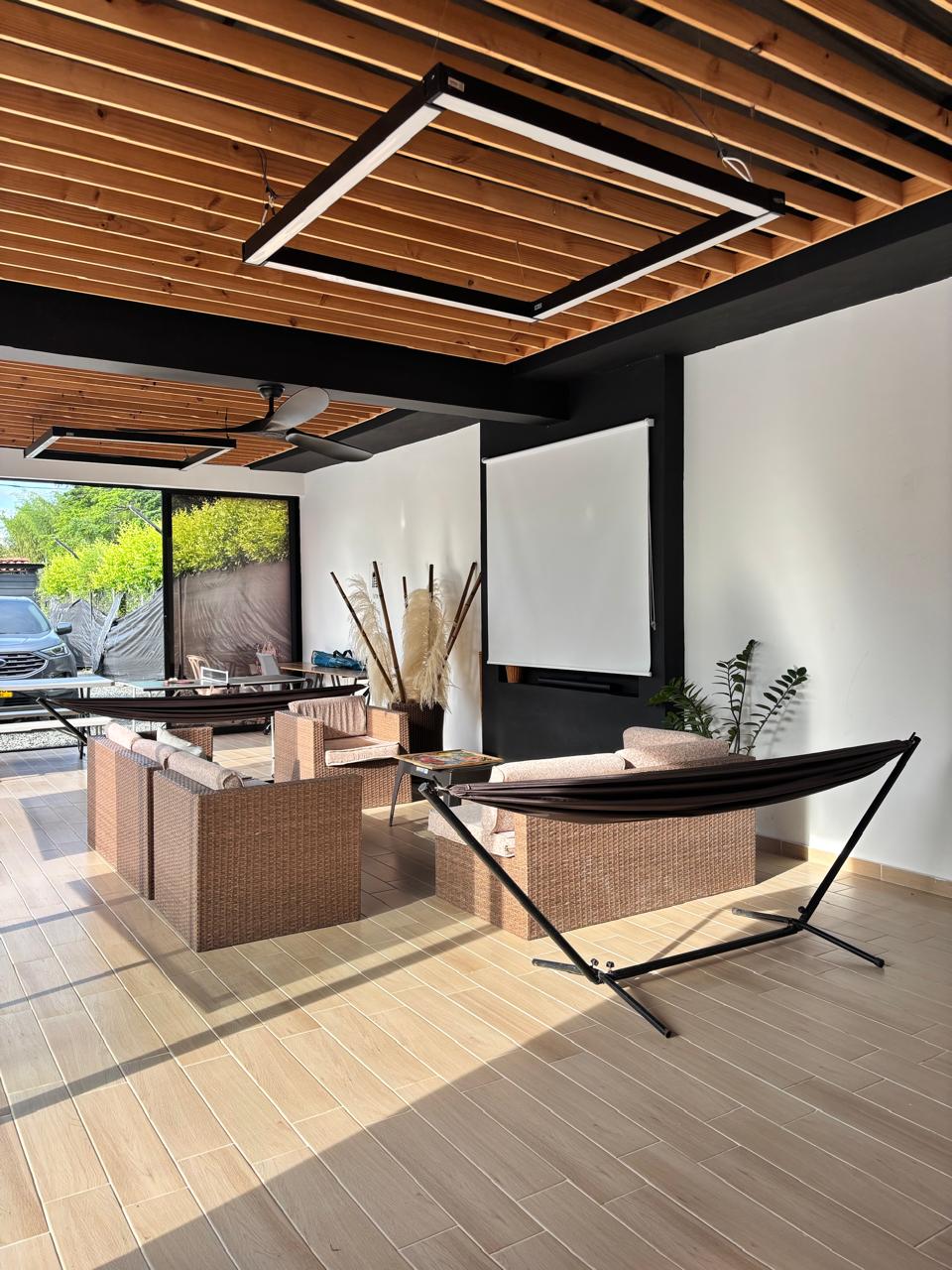 A modern lounge area features wicker seating arrangements complemented by a sleek, minimalist design. Two suspended light fixtures hang from the wooden ceiling, illuminating the space. A large screen is mounted on the wall, with natural light filtering through nearby windows.