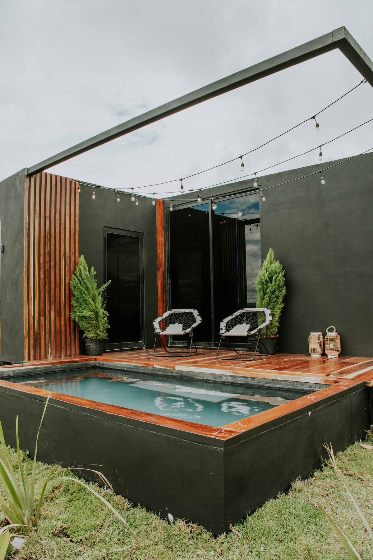 A sleek exterior showcases a black structure with wooden accents, featuring two seating chairs near a small plunge pool. Decorative elements include potted plants and soft lighting strung overhead, creating a modern and inviting outdoor space.