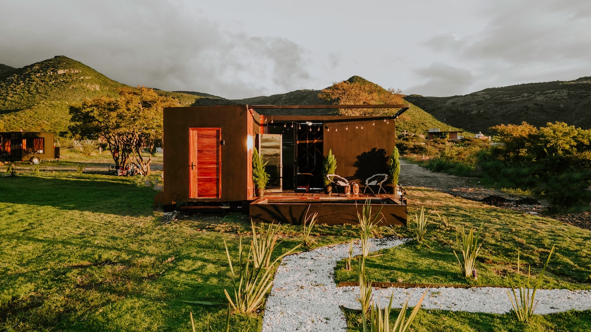 A modern Tiny House is positioned amidst verdant hills, framed by a pathway of gravel and greenery. The exterior is accented with wooden elements, featuring a warm door and large windows that offer views of the serene landscape.