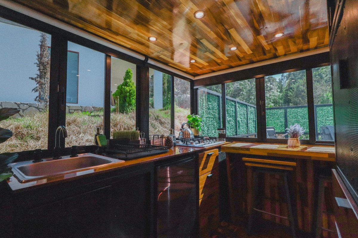 A compact kitchen is highlighted, featuring wooden ceilings and ample natural light from large windows. Modern appliances, including a stove and sink, are complemented by a cozy dining area with stools. Lush greenery is visible outside, enhancing the connection with nature.