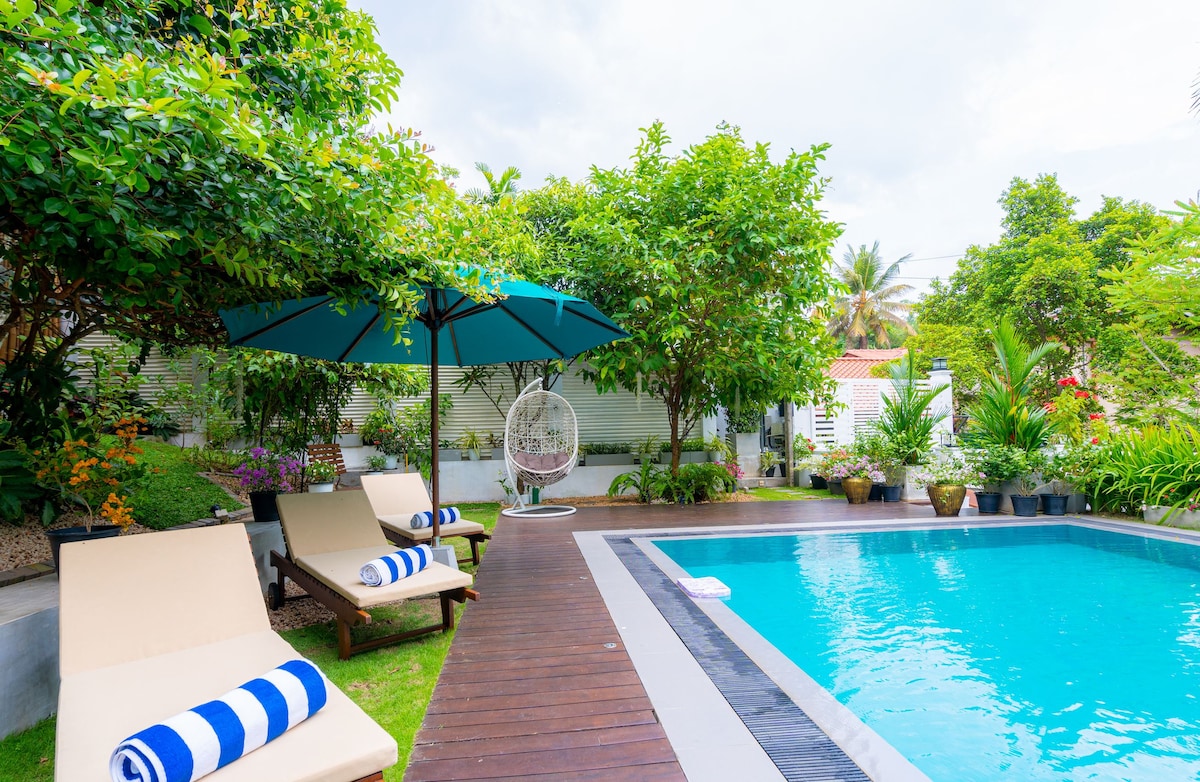 The outdoor area features a swimming pool surrounded by lush greenery. Sun beds are arranged with rolled blue towels, and a large umbrella provides shade. A hanging chair is positioned nearby, inviting relaxation amid the vibrant landscaping.