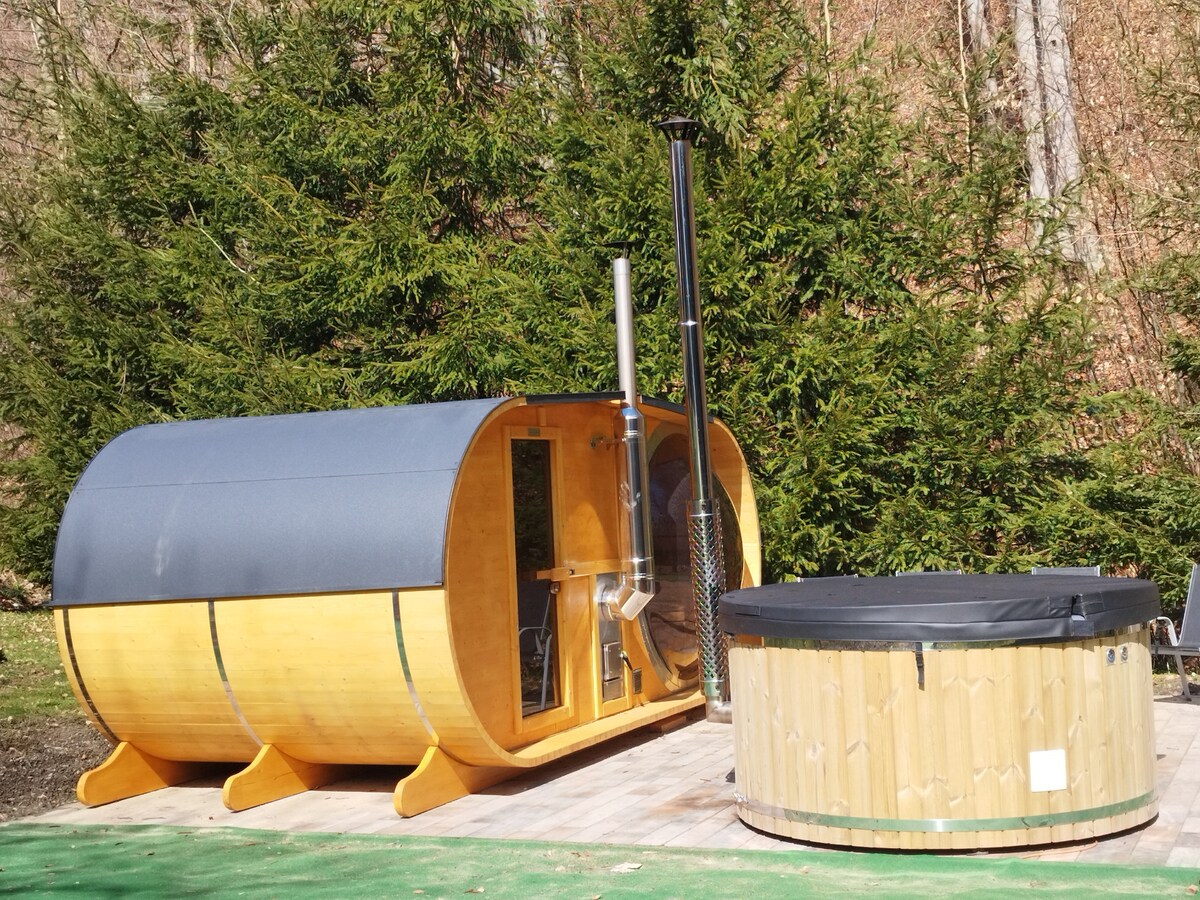 An outdoor wooden sauna is situated near a round wooden hot tub, both placed on a wooden deck. Surrounding the area are lush green trees, providing a natural backdrop. The sauna features a large window and a chimney, enhancing the rustic appeal.