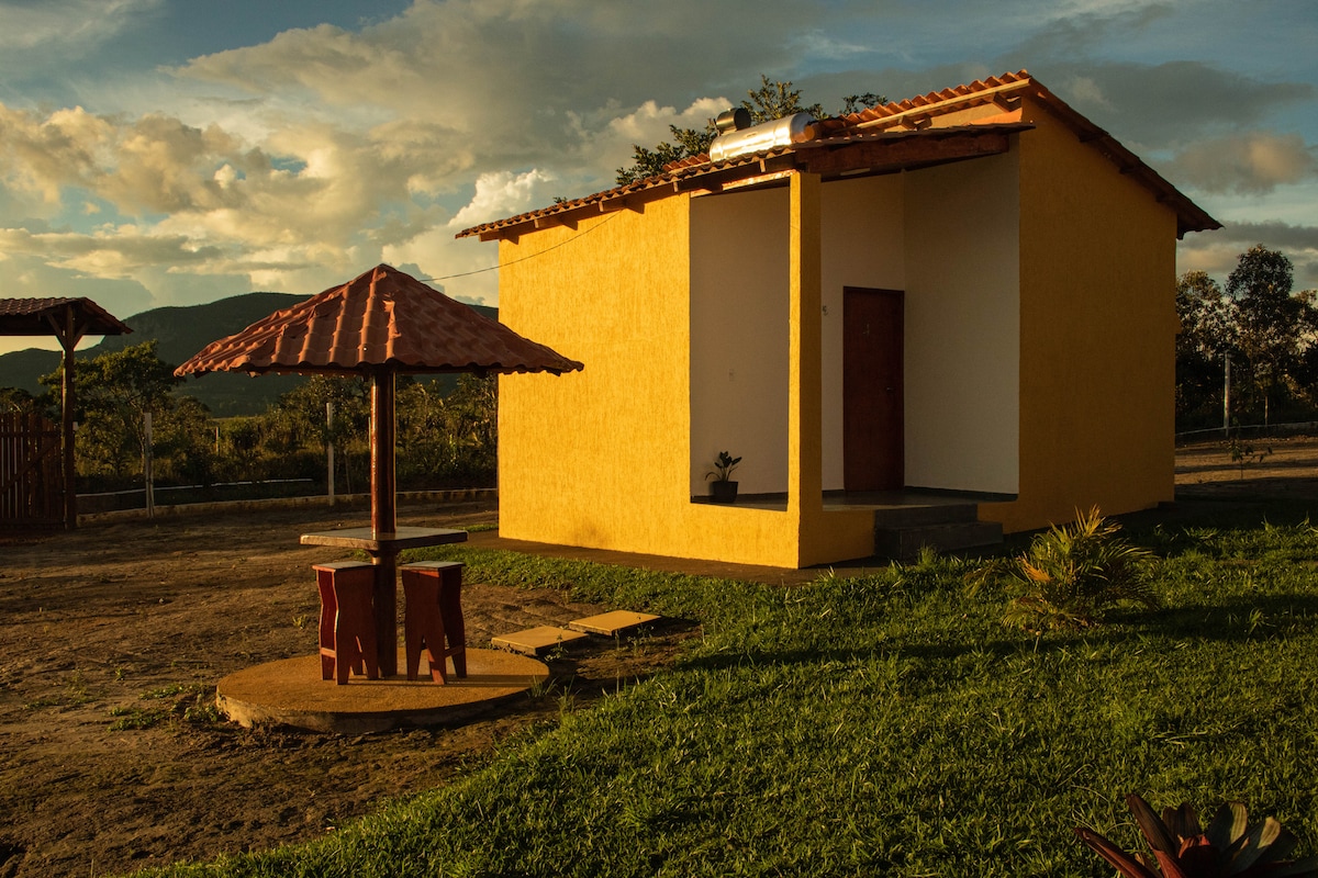 A single-story yellow chalet is set against a backdrop of hills. A shaded outdoor area features a small table with two chairs under a tile-roofed pergola. Lush green grass surrounds the building, which has a simple entrance and a small plant placed by the door.