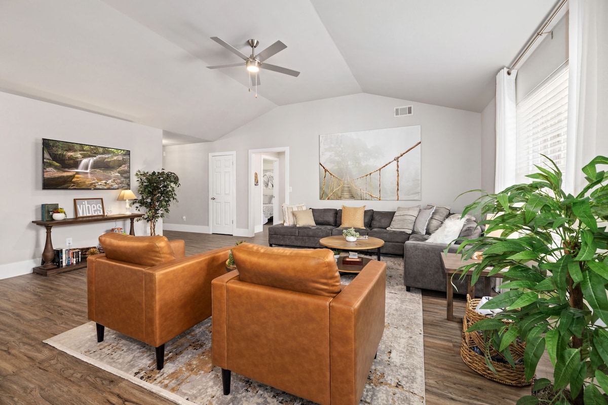 A spacious living room features a sectional sofa arranged around a coffee table. Two brown leather chairs provide additional seating. A television displaying a nature scene is mounted on the wall, with a ceiling fan overhead. Natural light enters through large windows, complemented by indoor plants.