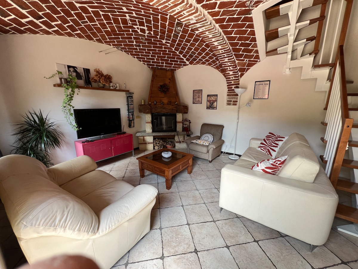 The living area features a distinct vaulted ceiling with exposed brickwork, creating a warm environment. A fireplace is positioned centrally, surrounded by comfortable seating including a large cream-colored sofa and armchairs. A wooden coffee table anchors the space, complemented by a television and decorative accents.