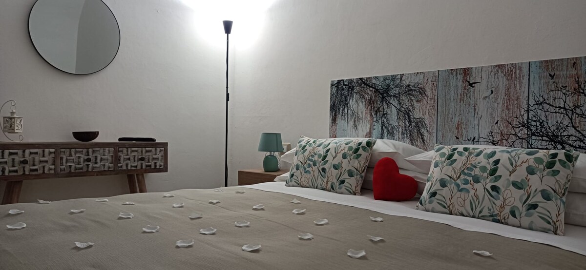 A cozy bedroom features a spacious bed adorned with decorative pillows and soft flower petals scattered across the beige comforter. A unique headboard with a nature motif is present, along with a lamp and a round mirror enhancing the calming atmosphere. Warm light illuminates the room.