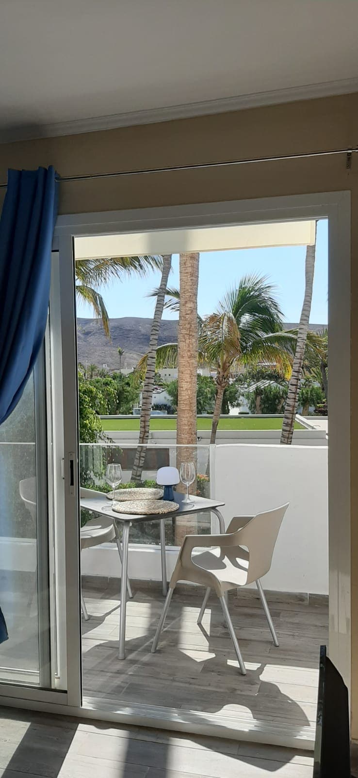 A sliding glass door leads to an outdoor balcony featuring a small round table set for two. Two chairs are positioned beside the table. Lush palm trees and green landscaping are visible in the background, creating a serene outdoor environment.
