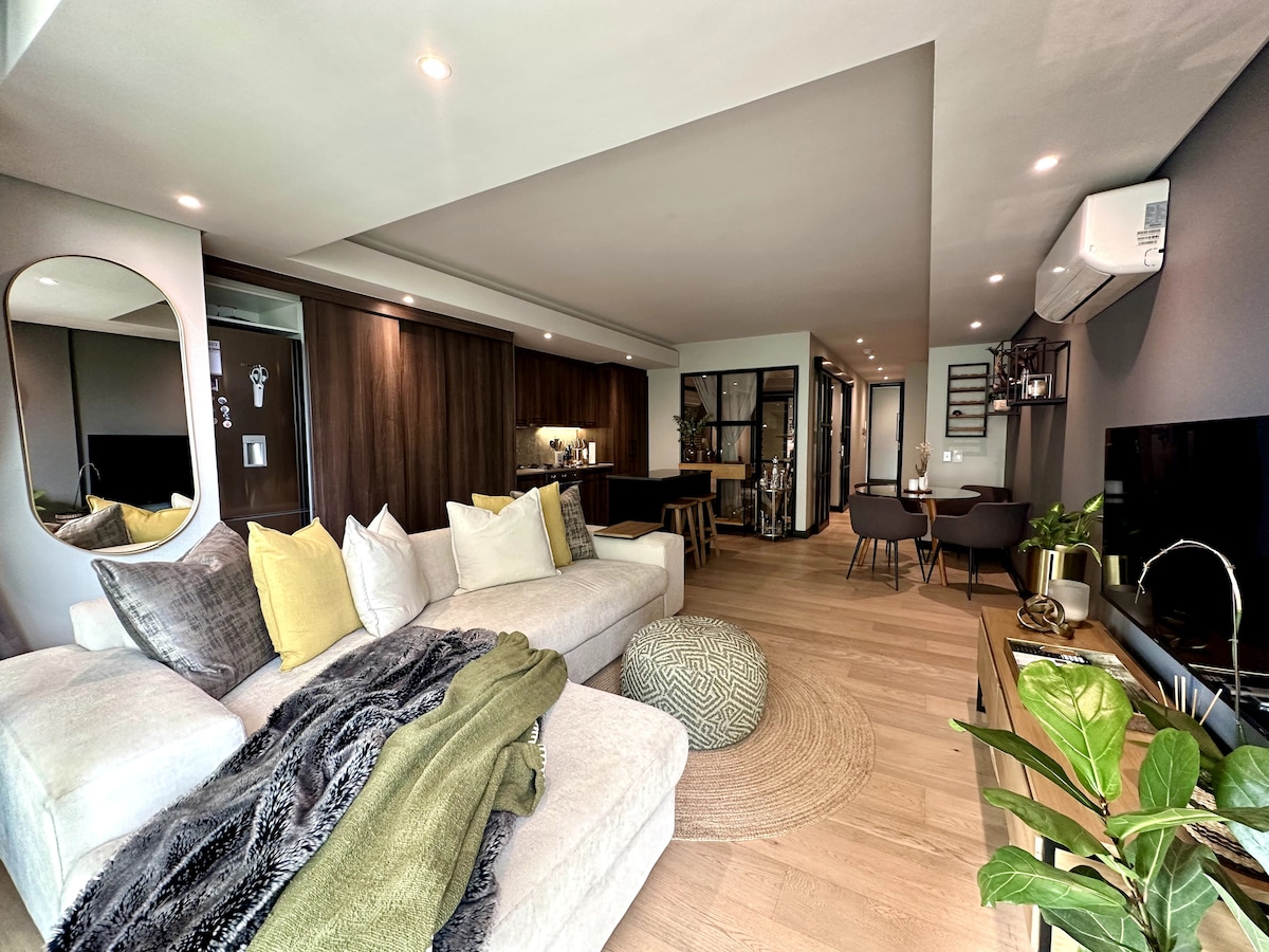 An inviting open-plan living area is highlighted by a spacious light-colored sectional sofa adorned with cushions and a soft throw. A round area rug sits beneath a coffee table, complemented by a sleek television unit. Natural light streams through windows, enhancing the modern design.