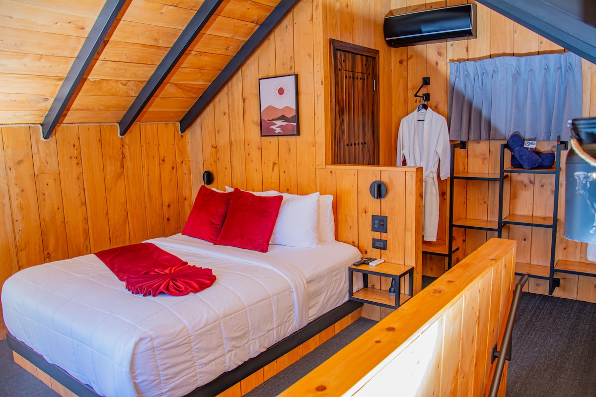 A cozy sleeping area features warm wooden walls and a sloped ceiling. A spacious bed is dressed in white linens with red accent pillows. A bedside table is positioned on one side, while a clothing rack and hanging robe are visible, enhancing the room's functionality.