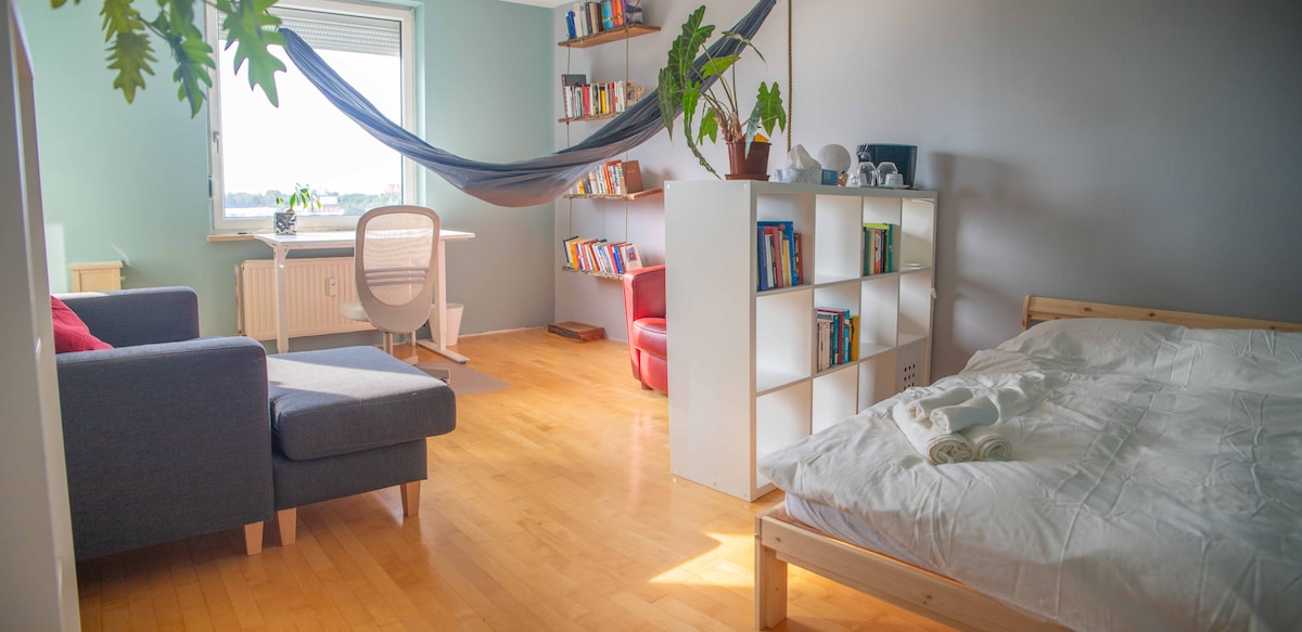 The room features a comfortable seating area with a couch and a reading chair, alongside a spacious bed with fresh linens. A hammock is suspended nearby, and a desk is positioned by the window. A bookshelf displays a collection of books, enhancing the serene environment.