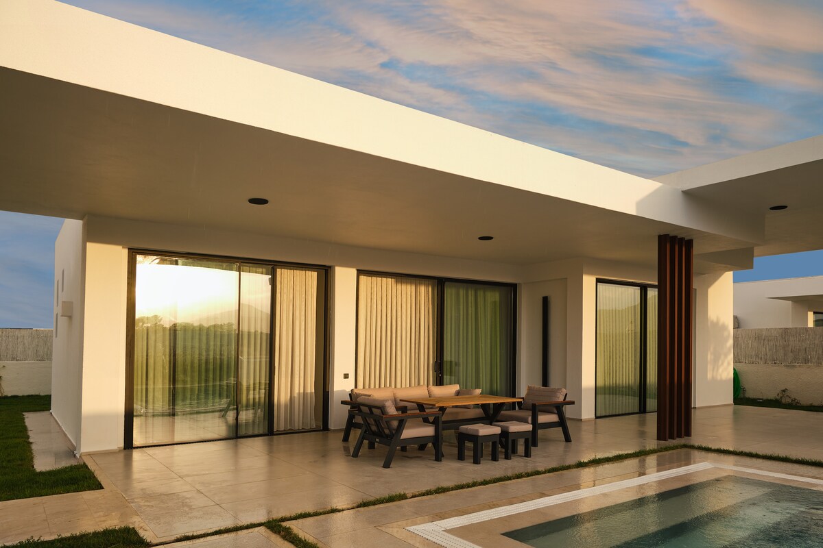 A modern villa exterior is showcased, featuring large sliding glass doors that connect indoor and outdoor spaces. A spacious patio area is furnished with a wooden dining table and comfortable seating. A private pool reflects the evening light, enhancing the serene atmosphere.