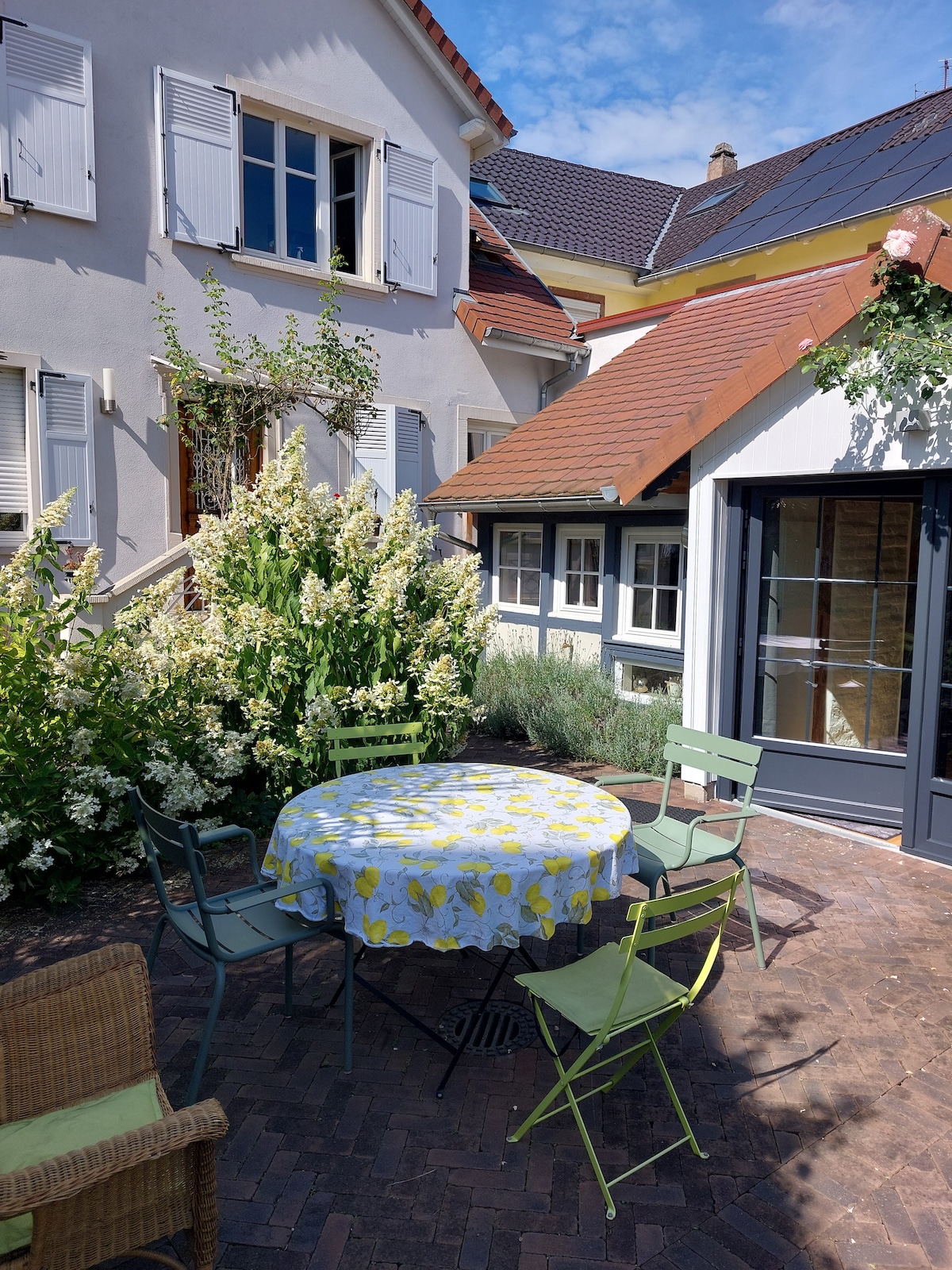 A charming outdoor space features a round table covered with a floral tablecloth, surrounded by four green chairs. Lush greenery and flowering plants provide a natural backdrop. The building's façade includes white shutters and a mix of brick and tiled surfaces.