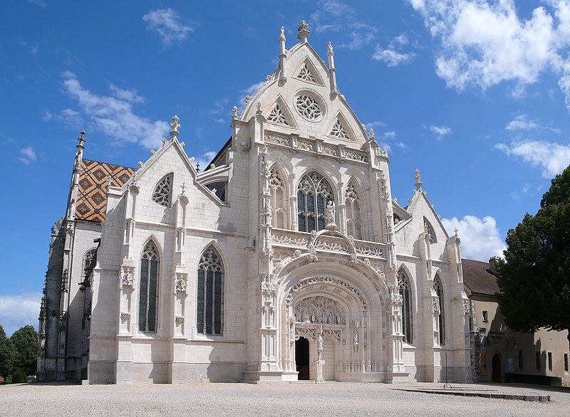 The exterior of a historic church is displayed, featuring intricate stone carvings and large stained-glass windows. A detailed facade is highlighted by pointed arches and ornate decoration, while a textured roof is visible in the background. A clear blue sky complements the structure.