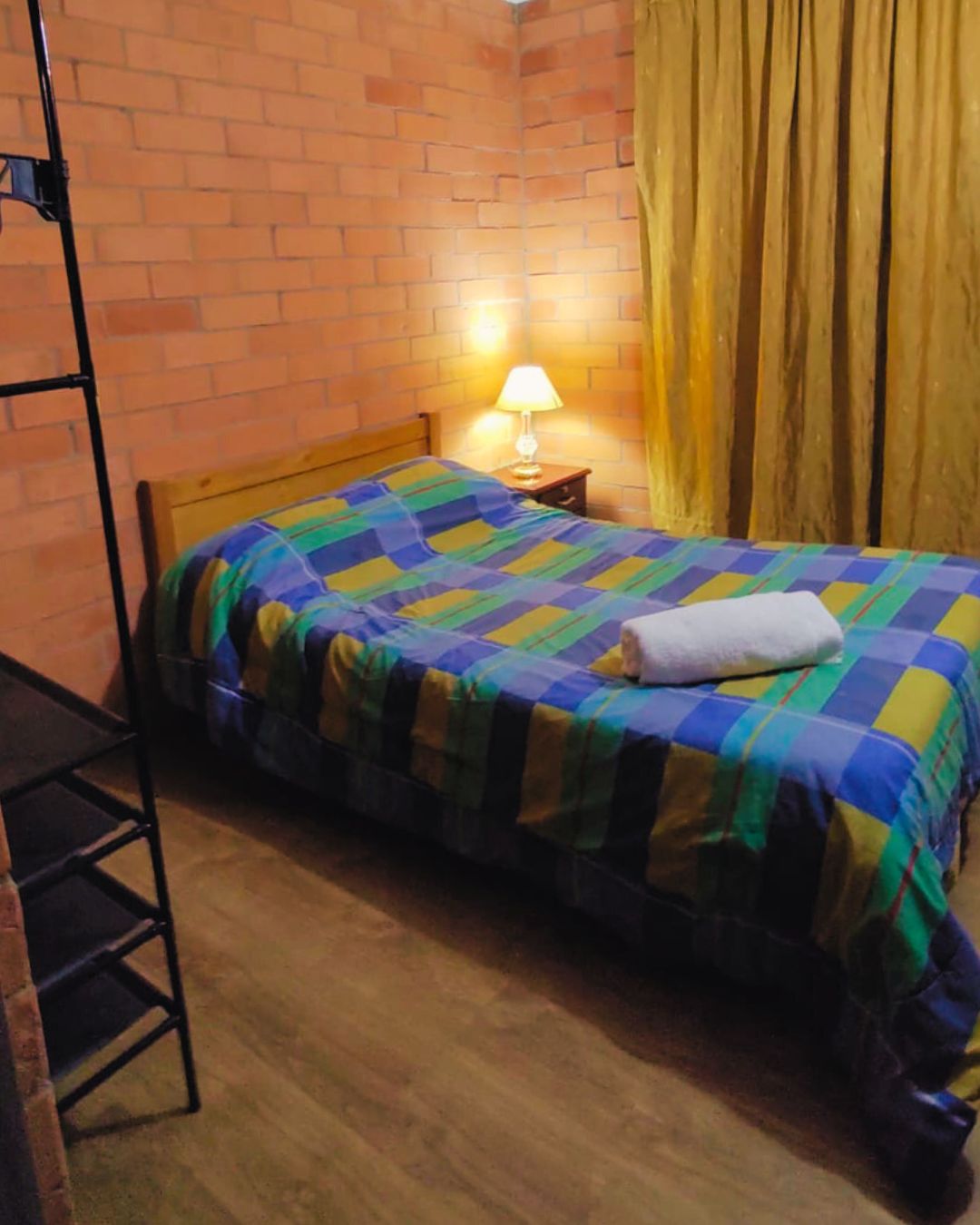 A cozy bedroom features a spacious bed adorned with a striped plaid blanket. A bedside lamp provides soft lighting next to a wooden nightstand. Warm, textured brick walls and soft golden curtains complement the relaxing ambiance.