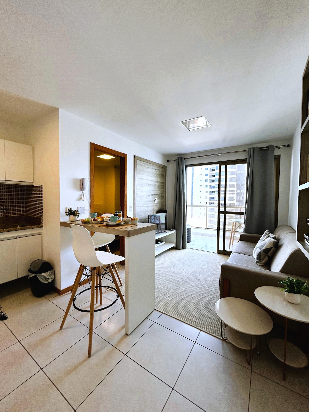 An open-plan living space features a cozy seating area with a sofa and a coffee table. A small kitchen with counter seating is visible. Natural light enters through large windows, while neutral tones create a calming atmosphere. The balcony offers an inviting outdoor space.