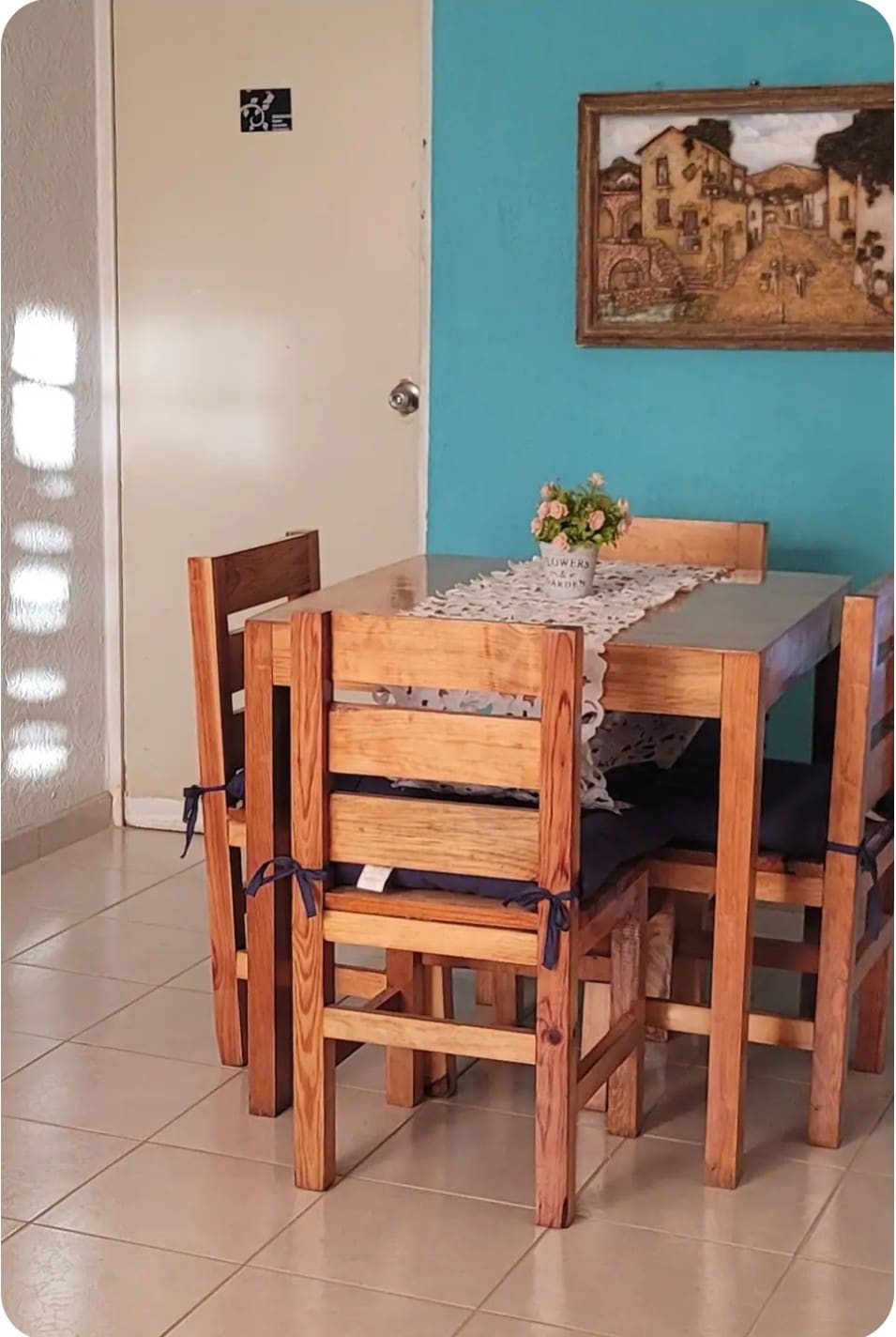 A wooden dining table is surrounded by four chairs, each with a cushion for comfort. The table features a lace runner and a small floral arrangement in the center. A wall is painted in a soothing turquoise, while a framed artwork is displayed nearby.