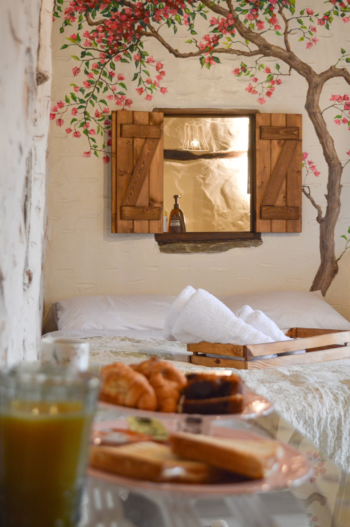 A cozy sleeping area features a spacious bed adorned with soft bedding. A decorative mural showcases blooming branches beside a framed mirror. A wooden tray presents a selection of breakfast items, accompanied by a glass of juice, adding a welcoming touch.