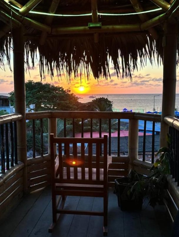 A bamboo gazebo offers a relaxing view of the sunset over the ocean. A wooden chair is positioned on the balcony, inviting quiet moments while enjoying the coastal scenery. Lush greenery can be seen nearby, enhancing the natural setting.