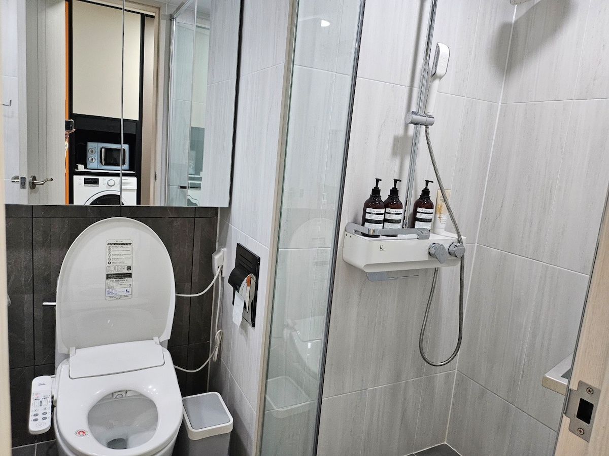 A modern bathroom is depicted, featuring a shower with a glass partition, a sink with toiletries displayed, and a toilet equipped with a bidet function. Neutral tones and sleek finishes create a clean and fresh environment, with a glimpse of a washing machine visible in the background.