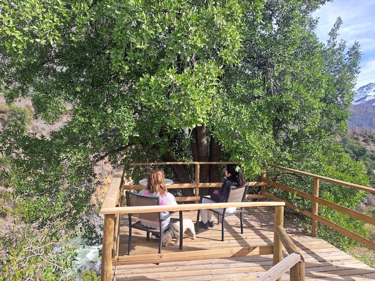 A wooden deck is nestled under a large green tree, providing a serene spot to relax. Two individuals are seated on comfortable chairs, enjoying the natural surroundings. The deck offers a view of the flowing water below and the distant mountains.