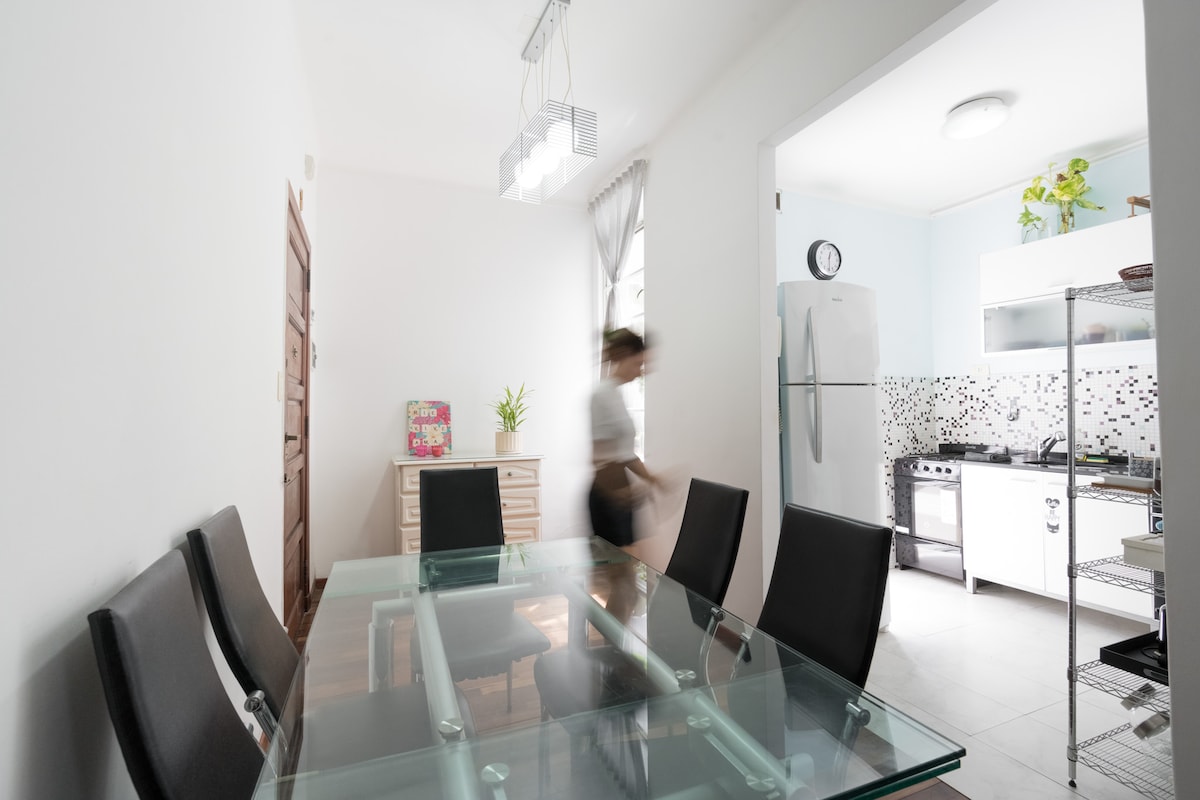 A modern dining area showcases a glass table surrounded by six black chairs. A side cabinet is positioned against the wall, with green plants decorating the space. The kitchen, visible in the background, features white cabinetry and a spacious refrigerator.