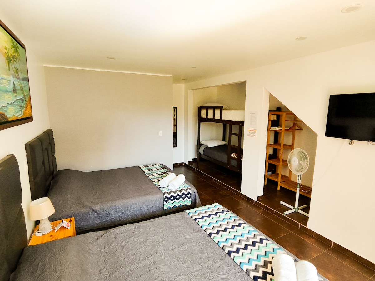 This spacious guest room features two large beds covered in gray linens. A wall-mounted television is visible, alongside a small fan. A bunk bed is positioned in the corner, with a decorative rug adding a touch of color to the tiled floor.