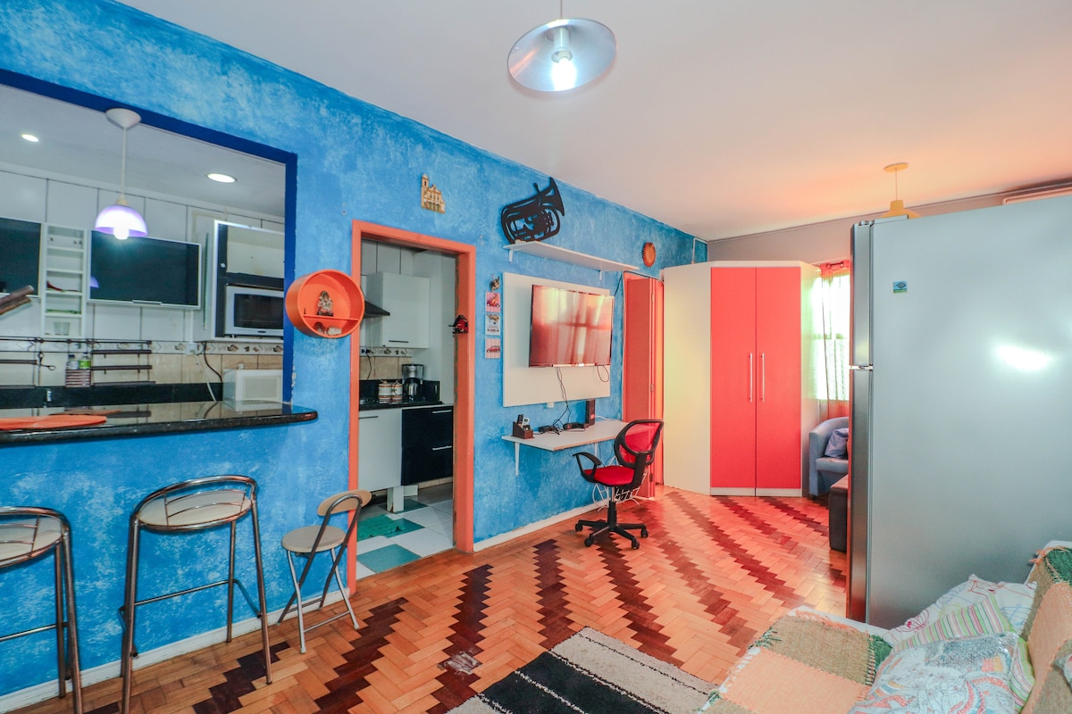 A functional studio space features a bright blue accent wall. A small dining area with two bar stools is present alongside a compact kitchen area with modern appliances. A seating area contains a chair facing a mounted TV, with decorative elements adding character to the room.