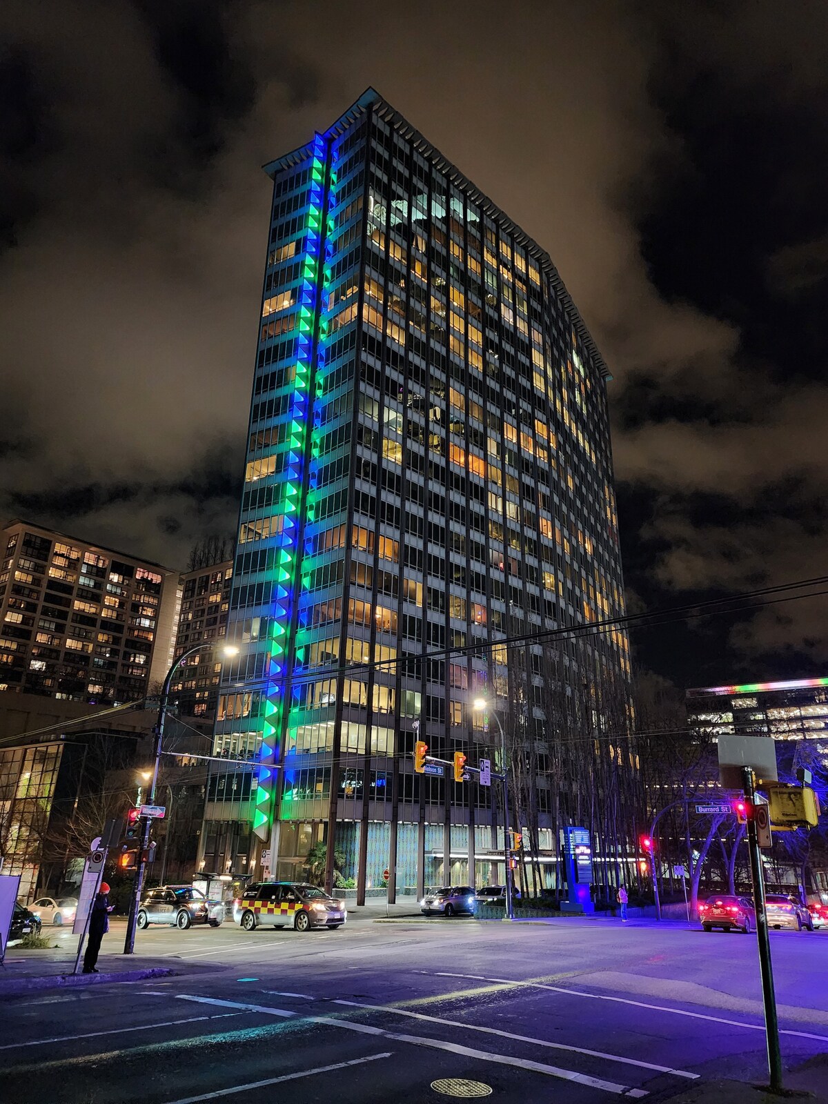 An urban high-rise building is illuminated at night, showcasing a vibrant blue and green glow along its sides. The expansive glass windows reflect the city lights, while surrounding streets are bustling with vehicles and pedestrians.