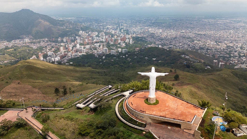 An expansive view of the city is presented, featuring urban buildings nestled against green hills. The prominent Christ the Redeemer statue overlooks the landscape, set on a well-maintained plaza. Rolling mountains serve as a backdrop, while the sky appears cloudy and dynamic.