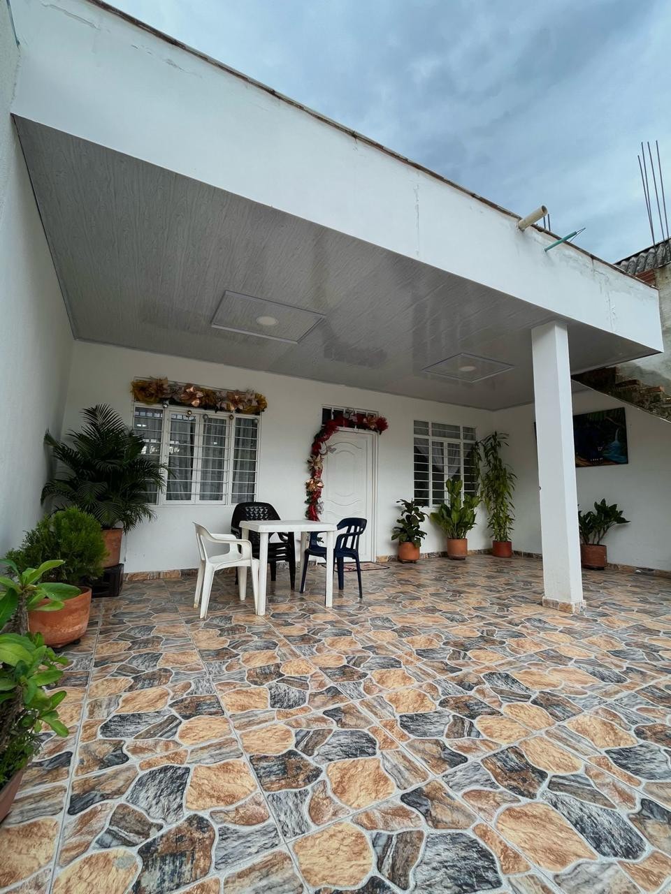 An outdoor patio area is outlined by potted plants, featuring a tiled floor in a natural stone pattern. A small table with four chairs provides a space for relaxation. The entrance is adorned with decorative elements, and large windows allow natural light to flow into the space.