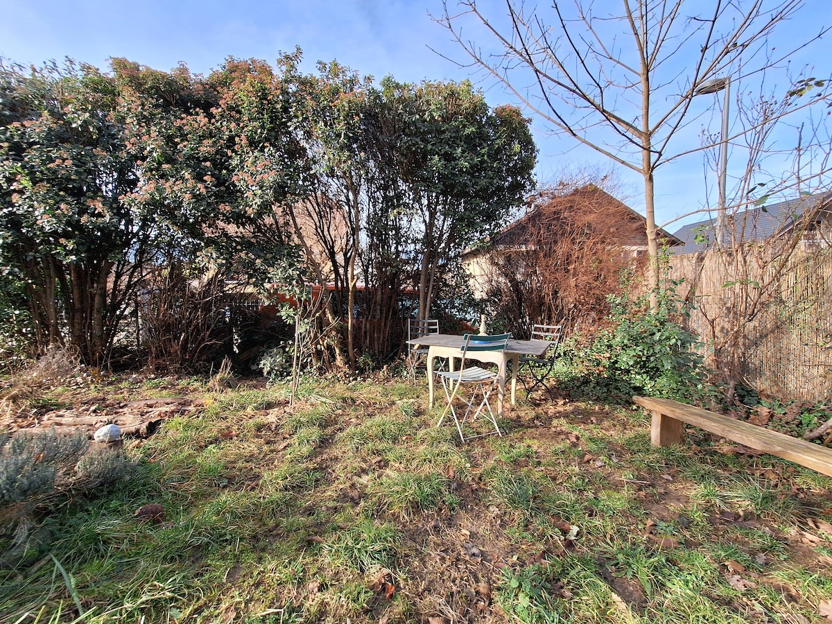 A private garden area is shown, featuring a small table with two chairs, surrounded by greenery and shrubs. The ground is covered in grass and scattered fallen leaves, with sunlight filtering through the trees, creating a serene outdoor setting.