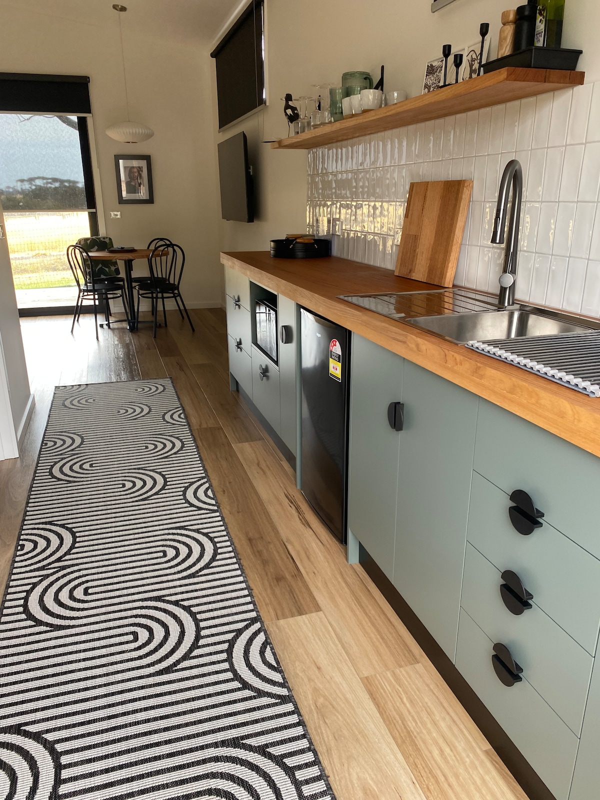 The kitchenette features light green cabinetry and a wooden countertop, with essential appliances including a refrigerator and portable induction cooktop. A black and white patterned runner adds a stylish touch to the floor. A dining area with black chairs is visible in the background.
