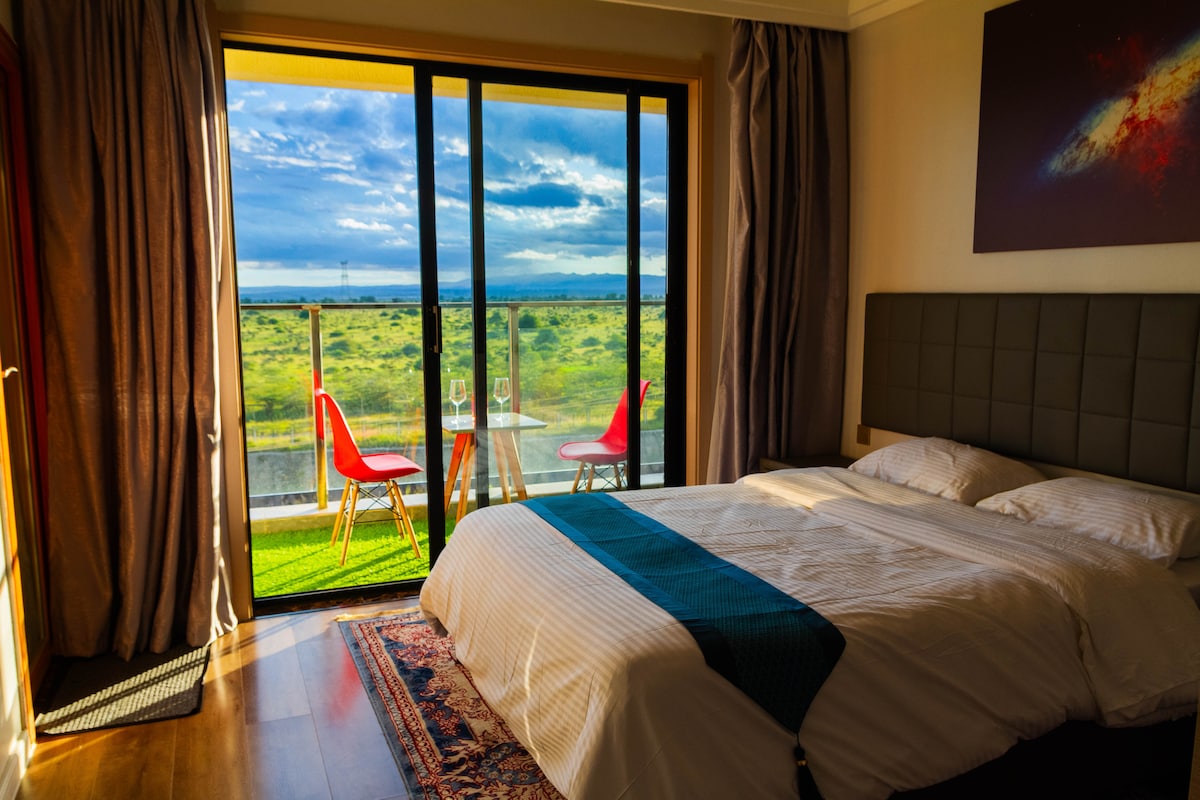 A bright bedroom features a spacious bed with crisp linens and a large window offering views of the landscape. Two red chairs and a small table are situated on the balcony, inviting relaxation amidst the scenic surroundings.
