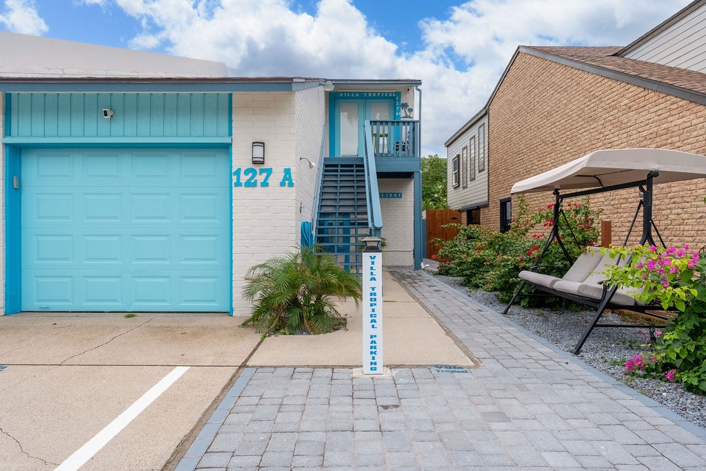 The entrance to the Townhome is framed by a light blue garage door and steps leading to the front door. A spacious driveway offers parking space, while a swing seat is positioned beside colorful landscaping with flowers and greenery. Cloudy skies provide a soothing backdrop.