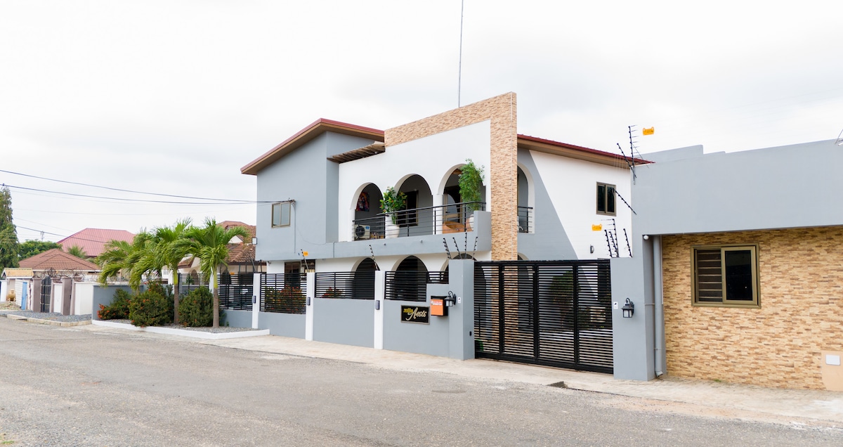 A modern two-story building is presented from the street view, featuring a combination of grey and beige exterior. Arched balconies and vibrant plants are visible, contributing to the overall aesthetic. The gated entrance provides a sense of security, while the surrounding area is characterized by open space.