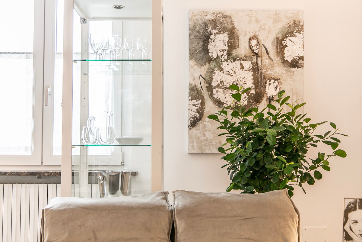A modern living area features a comfortable beige sofa, complemented by a decorative green plant. A glass cabinet displays elegant glassware, while a wall art piece with soft tones adds visual interest. Natural light illuminates the space through large windows.