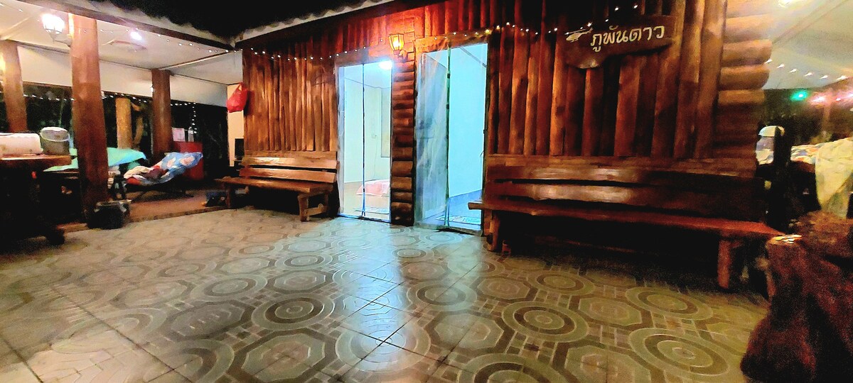 Wooden benches are positioned along the walls of a spacious indoor area, featuring intricate tile flooring. Large glass doors provide access to the outdoors, while soft lighting creates a relaxed ambiance, suitable for gathering with friends and family.