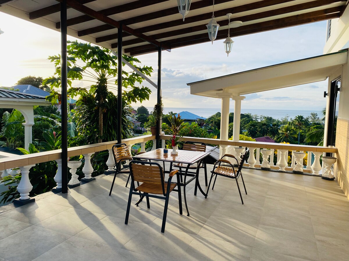 A spacious outdoor terrace is furnished with a wooden table surrounded by four chairs. The area is shaded by a roof, while lush greenery and distant ocean views can be seen in the background, creating a welcoming space for relaxation.