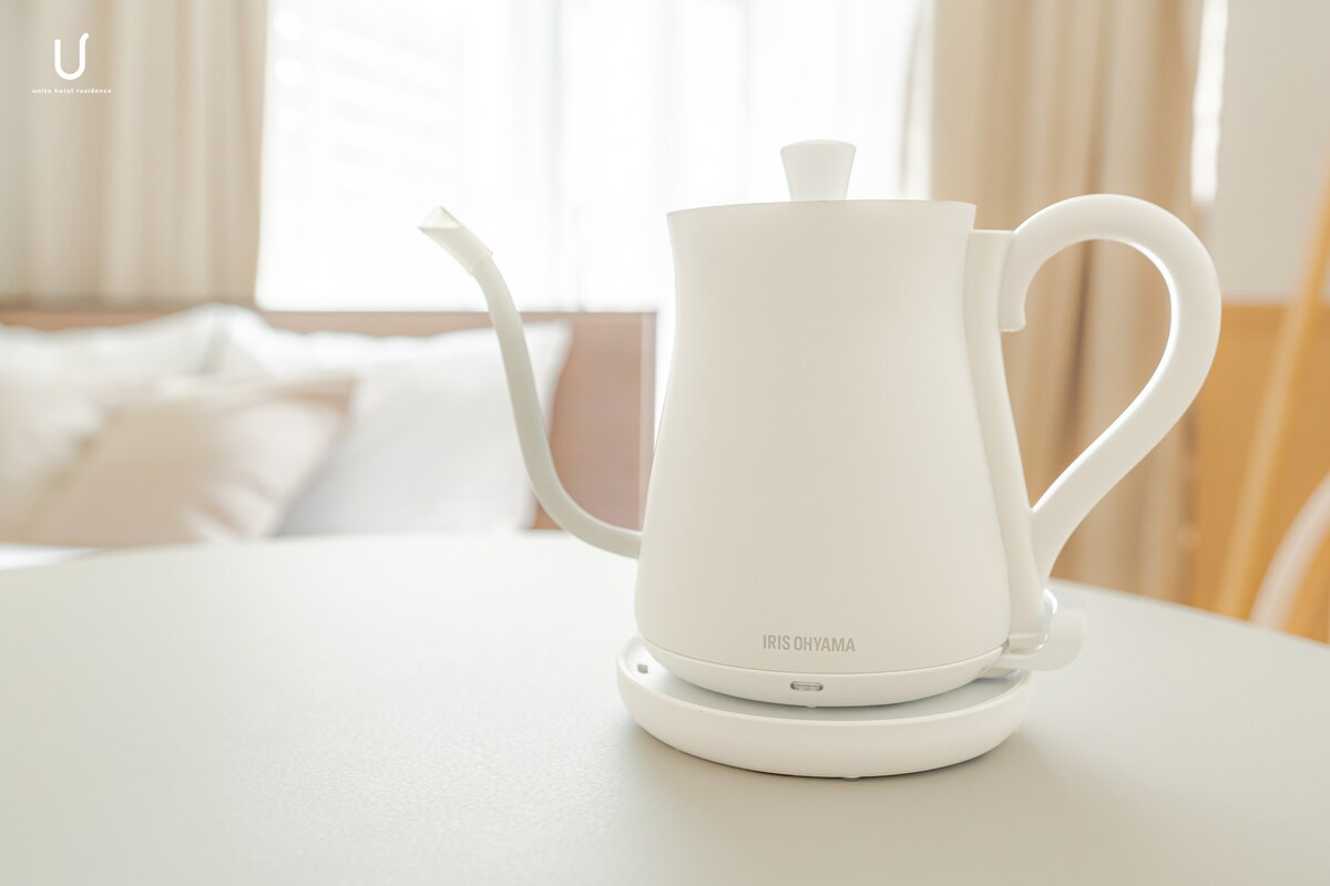 A sleek, white electric kettle is positioned on a minimalist base atop a round table. The kettle features a curved spout and an easy-grip handle. Soft, natural light filters through the nearby window, adding a calm ambiance to the setting.